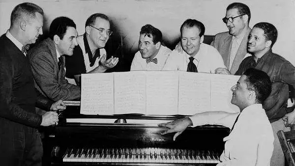 Benny Goodman (third from left) with some of his former musicians, seated around piano left to right: Vernon Brown, George Auld, Gene Krupa, Clint Neagley, Ziggy Elman, Israel Crosby and Teddy Wilson (at piano), 1952. World Telegram & Sun photo by Fred Palumbo. Library of Congress Prints and Photographs Division. Public domain.